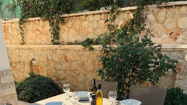 Mesa de comedor al aire libre con platos, vasos y una botella, rodeada de sillas y una pared de piedra con vegetación.