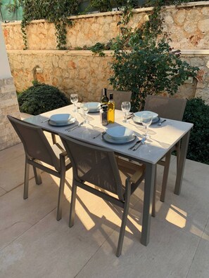 An outdoor dining table set with plates, glasses, and a bottle, surrounded by chairs and a stone wall with greenery. - Sylvana Apartment- a little piece of Kalkan heaven (Kalkan)