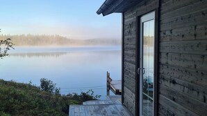 A wooden cabin with a glass door, a deck, and a view of a calm lake with a forest in the background.