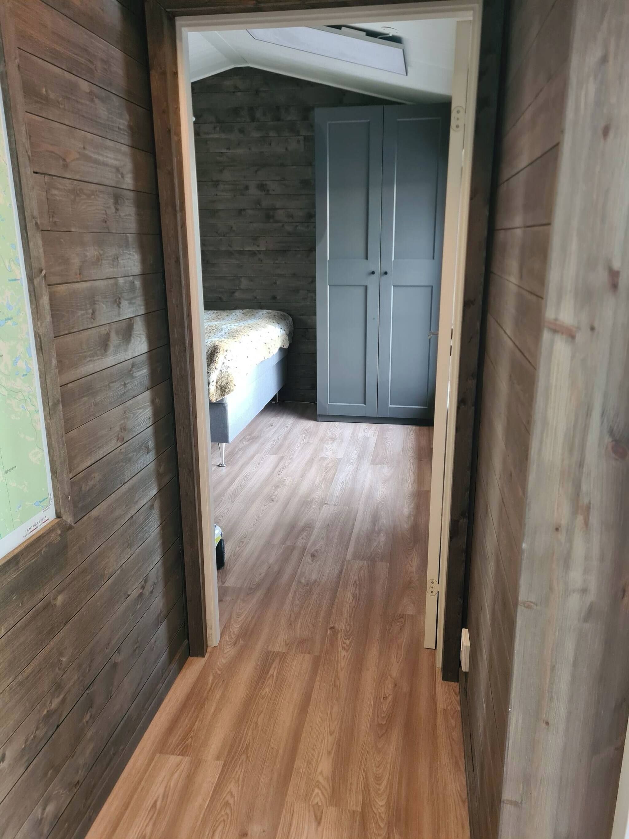A narrow hallway with wooden walls and a wooden floor leading to a bedroom with a bed and a wardrobe.