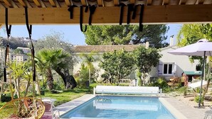 A covered outdoor area with a pool, a wooden table, a hanging chair, and a cat. - Provençal villa, swimming pool and wooded garden in a quiet location (Vence)