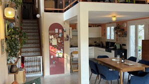 A spacious living area with a high ceiling, a dining table, blue chairs, a staircase leading to a loft, and a kitchen area. - Provençal villa, swimming pool and wooded garden in a quiet location (Vence)