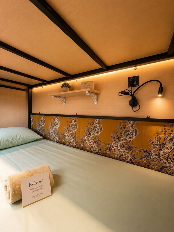 Una habitación de hotel con una cama, un letrero de bienvenida, un estante en la pared con un cartel, y una lámpara de pared.