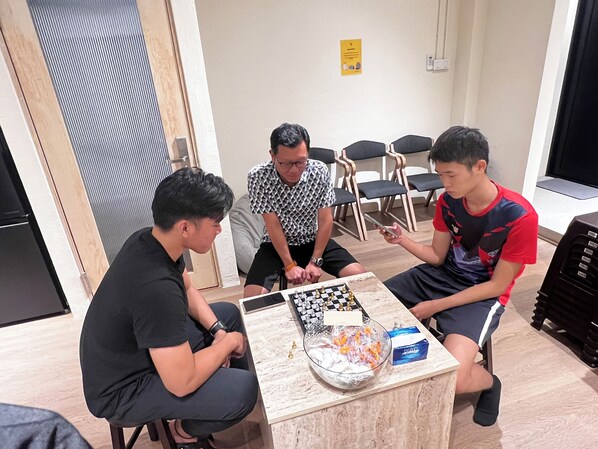 A group of people playing chess in a room with a wooden floor, a chessboard, and a bowl of snacks. - Urban Capsule by Faradale (Kuching)