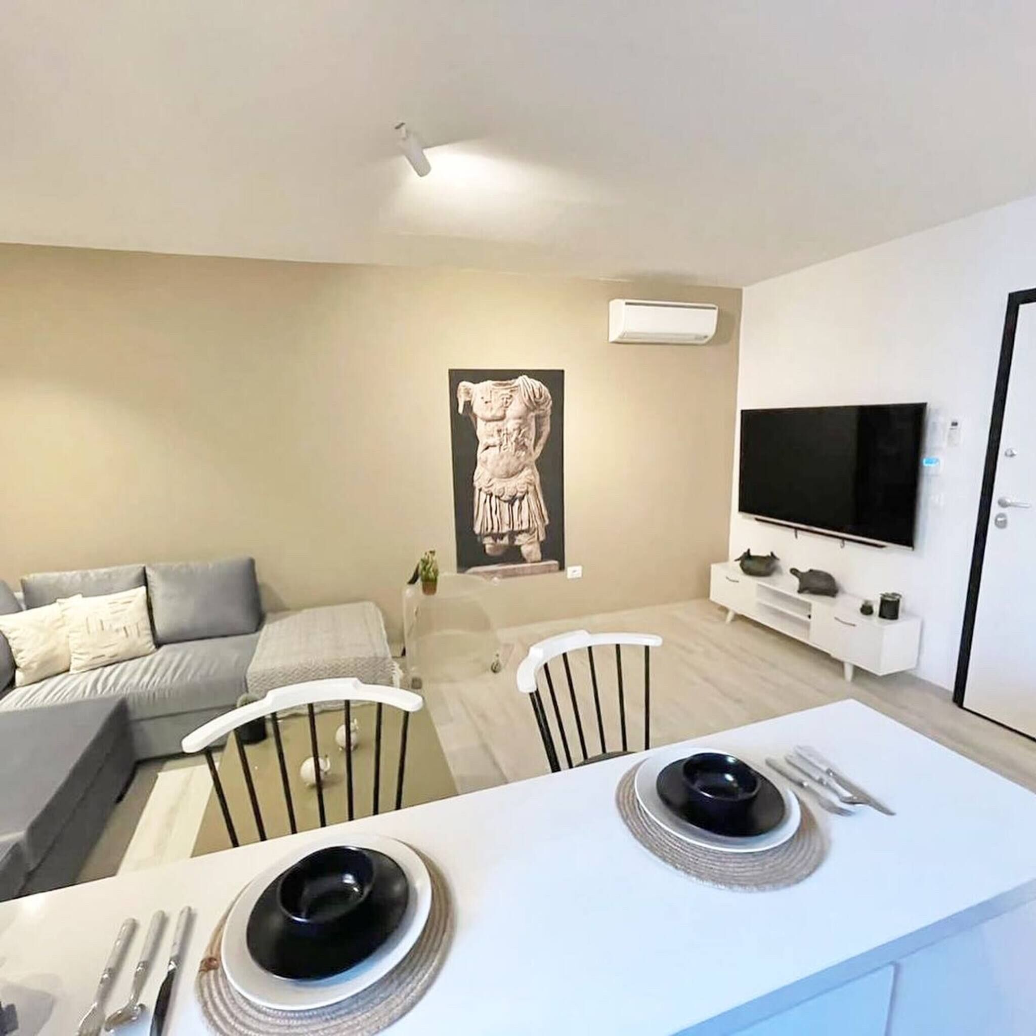 Un salon moderne avec un canapé gris, un coin repas doté d’une table blanche et un téléviseur fixé au mur.