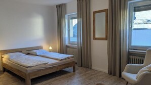 Ein Zimmer mit einem hölzernen Bett, einem Sessel, einem Tisch und einem Vorhangfenster.