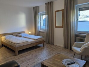 A room with a wooden bed, a chair, a table, and a window with curtains. - Holiday apartment Kuxborn in our guesthouse in the old town of Zell (Moselle) (Zell (Mosel))