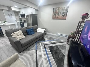A modern living room with a grey sofa, a flat-screen TV, and a kitchen in the background. - Cozy 1-bedroom  Suite in Saskatoon -Fully Furnished near Industrial Area/Shops (Saskatoon)