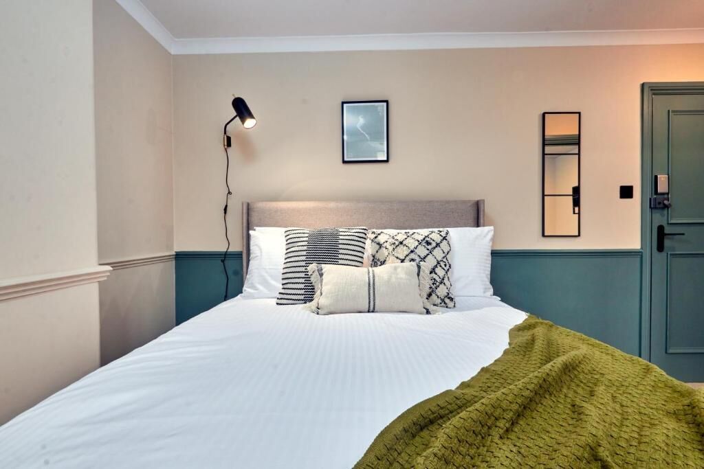 Een net opgemaakt bed met een groene deken, een bedlampje en een ingelijste foto aan de muur.