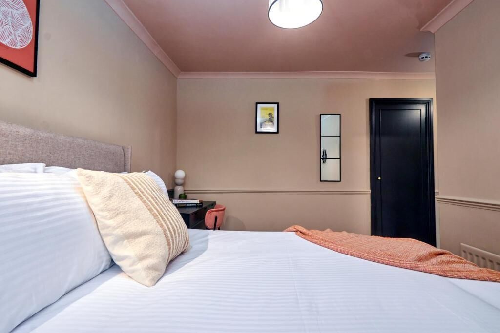 En pænt redt seng med puder, et sengebord med en lampe, et billede i ramme på væggen og en dør.