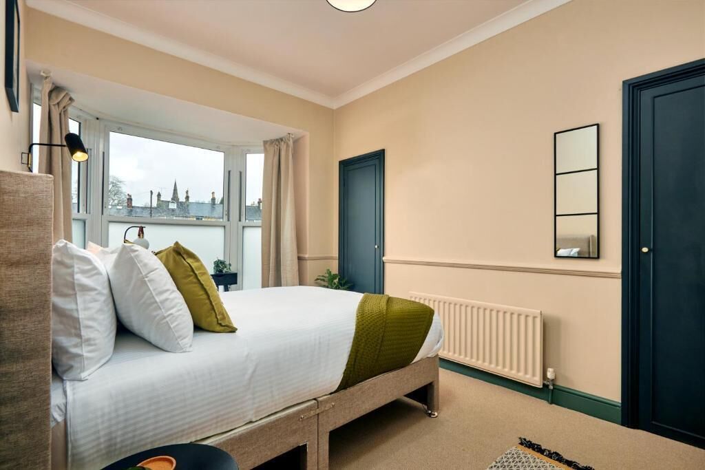 A bedroom with a bed, a window, a radiator, a door, and a mirror.