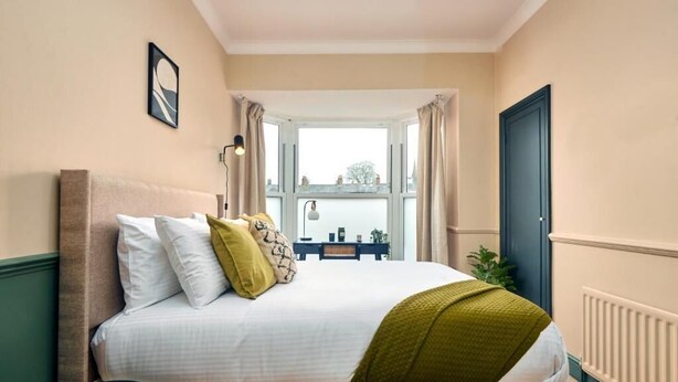 A bedroom with a bed, a nightstand, a lamp, a window with curtains, and a radiator. - Settle Winchester (Winchester)