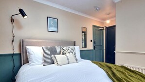 A neatly made bed with white bedding and patterned pillows, a black desk lamp, a framed picture on the wall, and a door in the background. - Settle Winchester (Winchester)