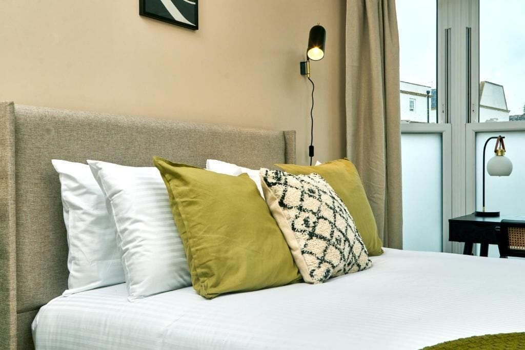 A bed with multiple pillows, a bedside lamp, and a window with a view of buildings.