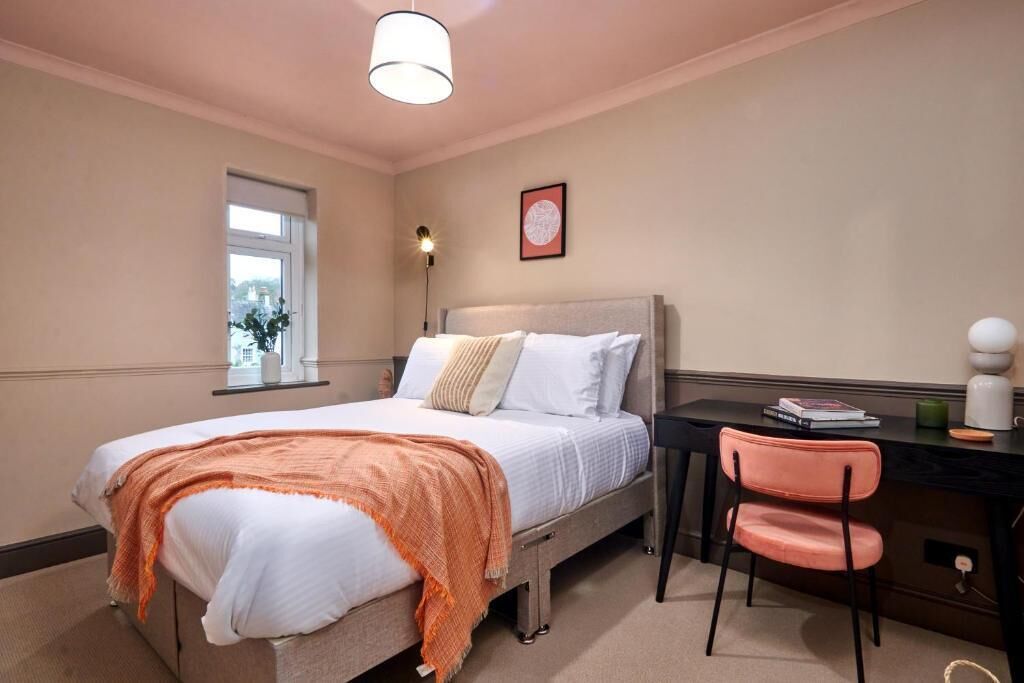 A bedroom with a bed, a desk, a chair, a window, and a wall-mounted light fixture.