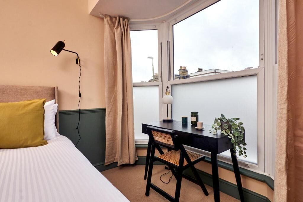 Een slaapkamer met een bed, een bureau, een stoel, een lamp en een uitzicht op gebouwen door een raam.