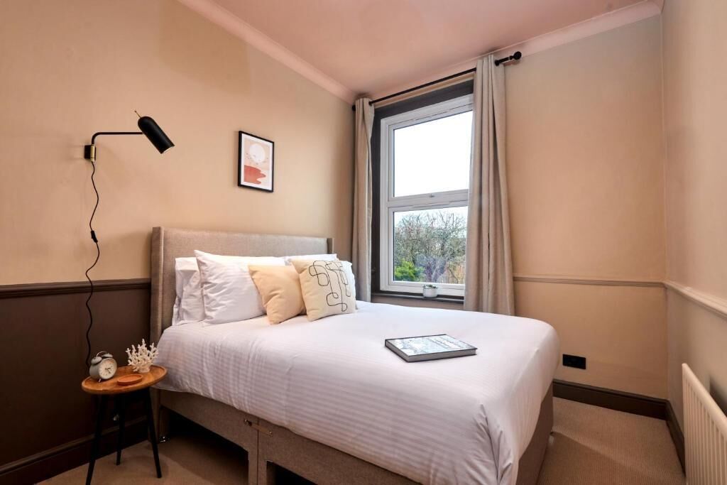 Een slaapkamer met een bed, een nachtkastje met een lamp, een lijst aan de muur en een raam met gordijnen.