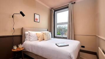 Een slaapkamer met een bed, een nachtkastje met een lamp, een lijst aan de muur en een raam met gordijnen.