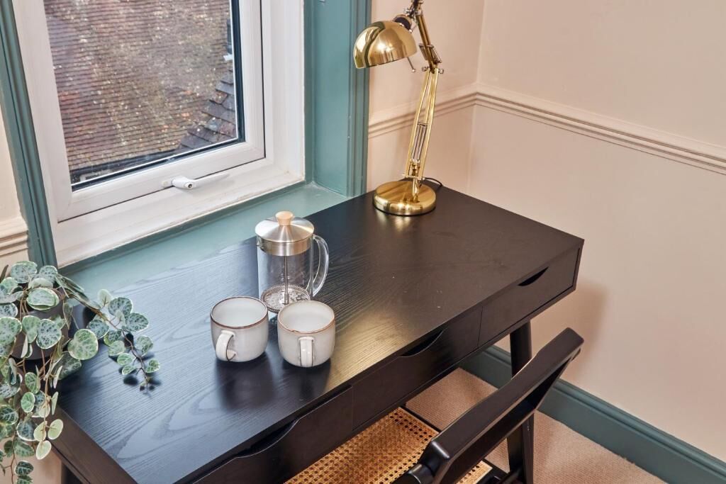 A dark wooden desk with a brass desk lamp, two white mugs, a glass container, and a potted plant.