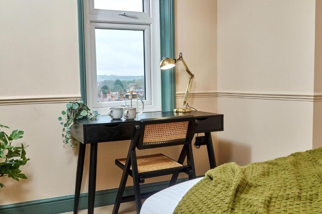 Une pièce comprenant un bureau, une chaise, une fenêtre, une lampe et offrant une vue sur le paysage urbain.
