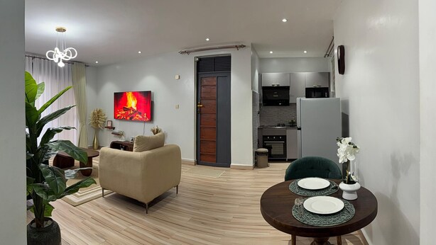 A modern living room with a sofa, a dining table set for two, a kitchen in the background, and a large TV mounted on the wall. - Luxurious 1 BR Apt in quaint Kampala with power backup. (Kampala)