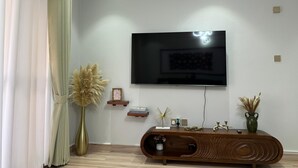 A modern living room with a flat-screen TV mounted on the wall, a wooden console table with decorative items, and a tall vase with dried plants. - Luxurious 1 BR Apt in quaint Kampala with power backup. (Kampala)