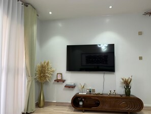 A modern living room with a flat-screen TV mounted on the wall, a wooden console table with decorative items, and a tall vase with dried plants. - Luxurious 1 BR Apt in quaint Kampala with power backup. (Kampala)