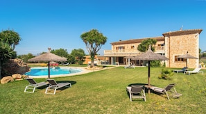 A poolside area with lounge chairs and umbrellas, a stone building with a balcony, and a clear blue sky.