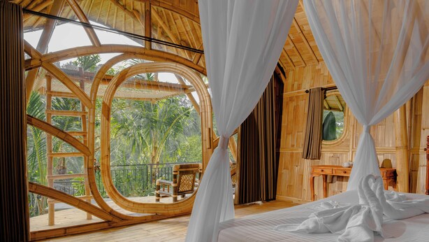 A room with a bed, a wooden floor, a large window with a view of greenery, and a canopy bed.