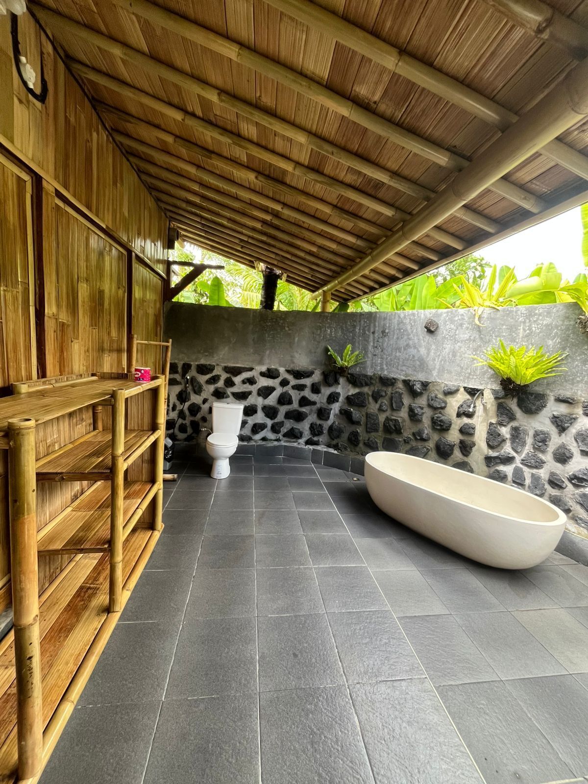 A bathroom with a stone wall, a toilet, a large bathtub, and a bamboo shelf.