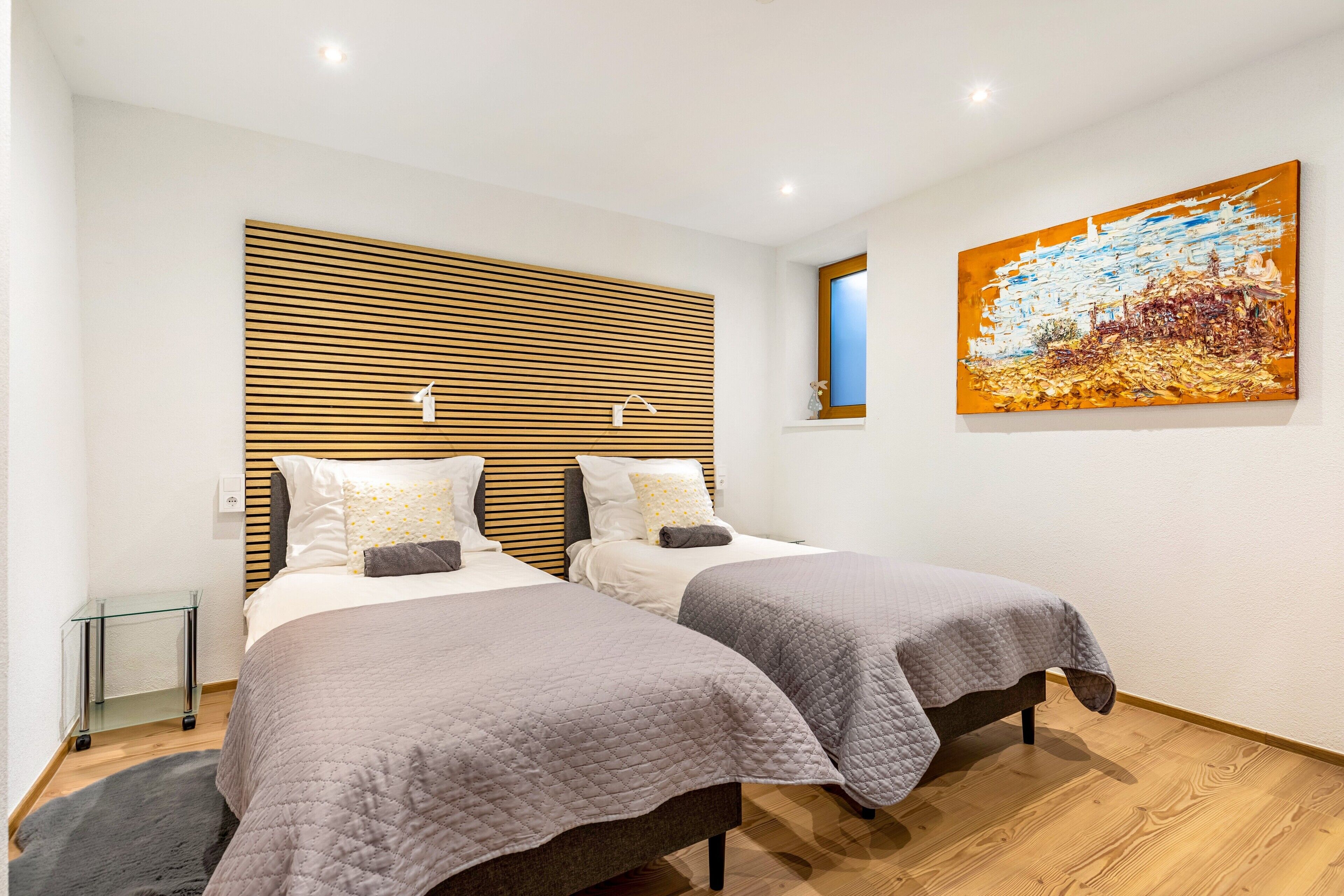 Une chambre d’hôtel moderne avec deux lits, une tête de lit en lattes de bois, une grande peinture abstraite et une petite fenêtre.