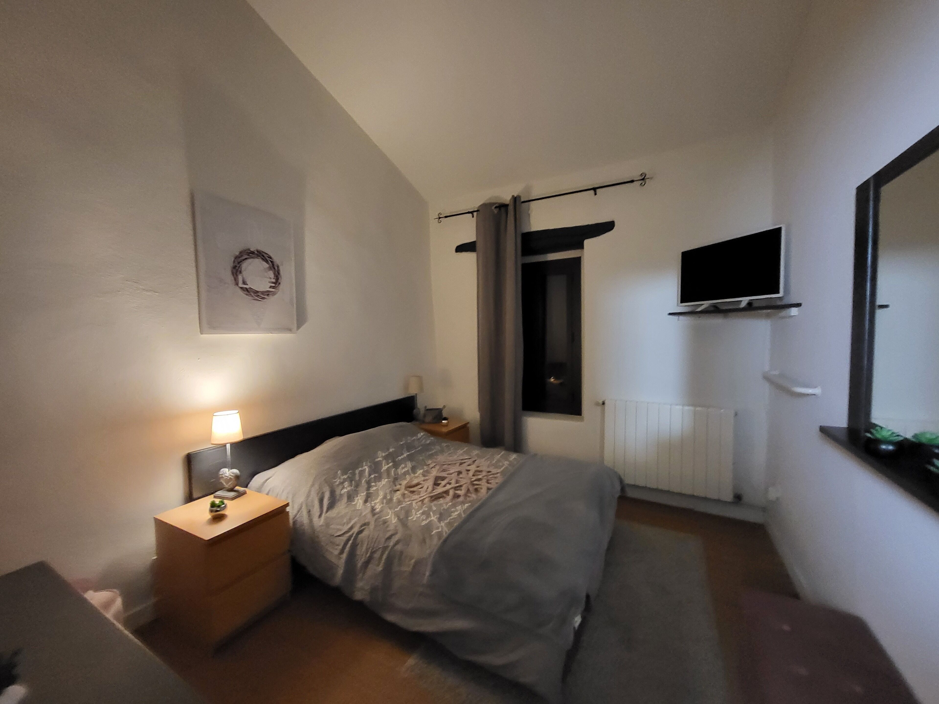 A bedroom with a bed, a nightstand, a wall-mounted TV, a radiator, and a window with curtains.