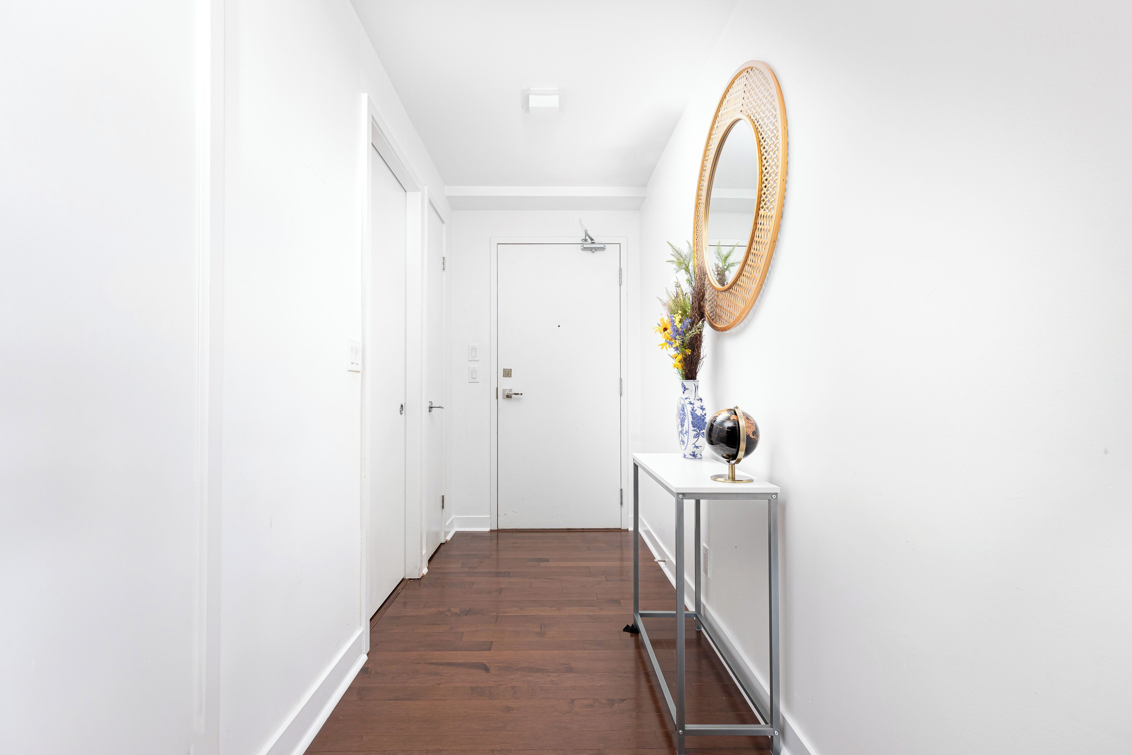 A hallway with white walls and wooden flooring, leading to a door. A small table with a vase and a globe is placed against the wall.