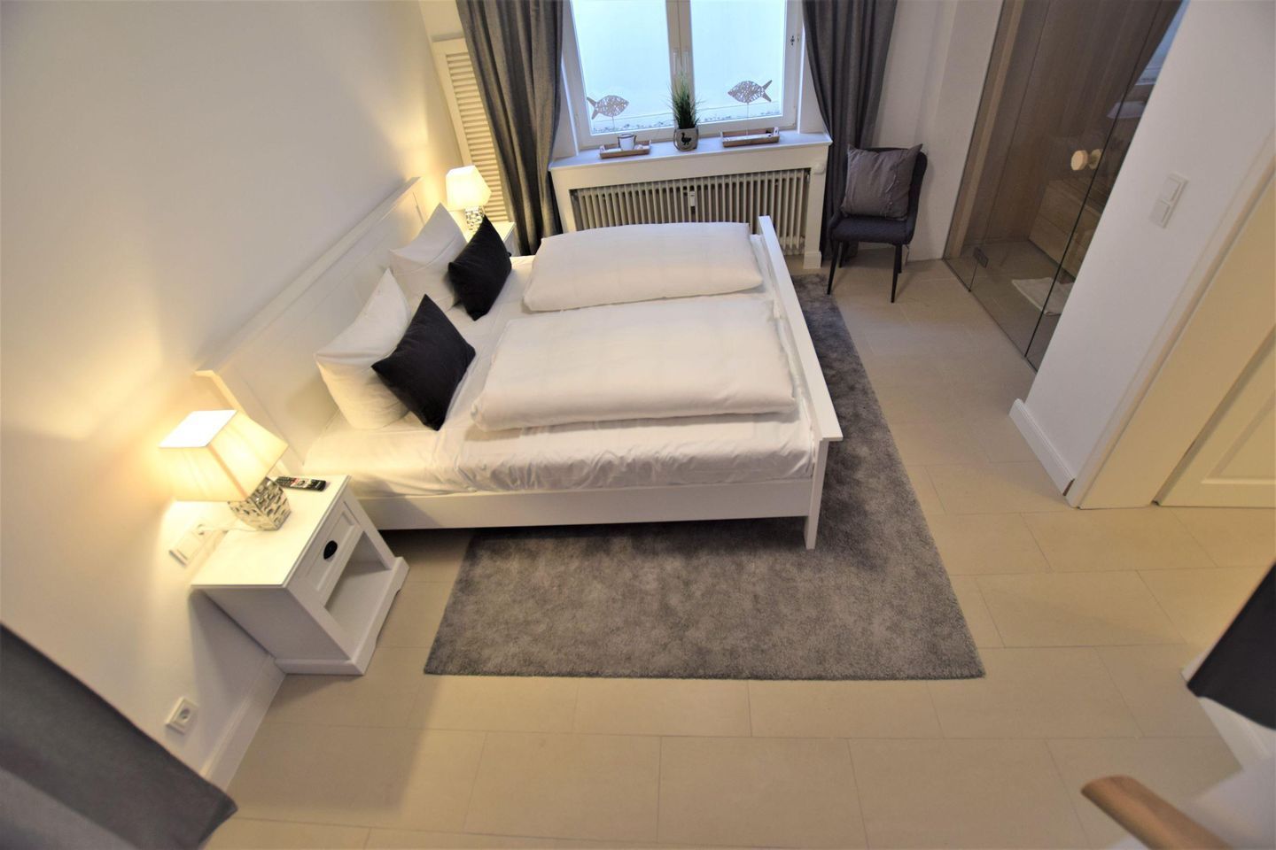 Ein Schlafzimmer mit einem Bett, Nachttischen, einer Lampe, einem Sessel, einem Fenster mit Vorhängen und einem Heizkörper.