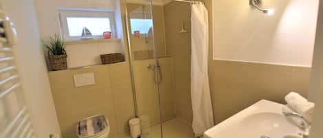 A bathroom with a glass shower enclosure, a toilet, a sink, and a window with a view.