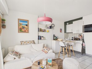 A modern living room with a white sofa, a coffee table, and a kitchen area with white cabinets and a pink pendant light. - Superb apartment for 2 with sea view (Saint-Gilles-Croix-de-Vie)