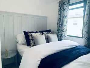 A neatly made bed with white and dark blue bedding, a small bedside table with a glass top, and a window with patterned curtains. - Hopton Holiday Village - Holiday Accommodation 12171 (Great Yarmouth)