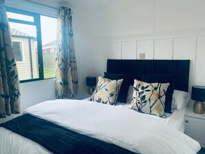 A bedroom with a bed, curtains, a nightstand, and a lamp. - Hopton Holiday Village - Holiday Accommodation 12171 (Great Yarmouth)