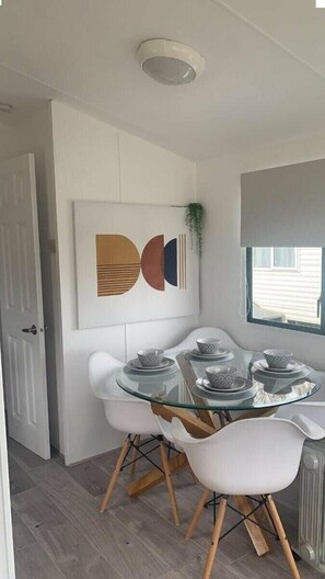A dining area with a glass table, white chairs, and a wall-mounted abstract art piece. - Hopton Holiday Village - Holiday Accommodation 12171 (Great Yarmouth)