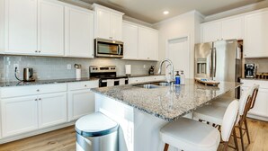 Une cuisine moderne avec des meubles blancs, des plans de travail en granit, des appareils électroménagers en acier inoxydable et des tabourets de bar.