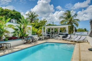 Ein Swimmingpool mit Liegestühlen, ein Grillplatz und eine überdachte Sitzgruppe, umgeben von Palmen.