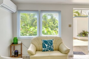 Ein Zimmer mit einem beigen Sofa, einer grünen Vase und einem weißen Kissen auf einem Holztischchen. Zwei Fenster mit Jalousien und eine Klimaanlage an der Wand sind vorhanden.