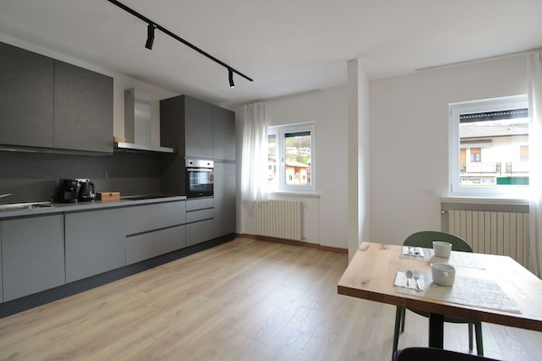 A modern kitchen with grey cabinets, a built-in oven, and a coffee maker. There's a dining area with a wooden table and chairs.