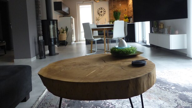 Ein modernes Wohnzimmer mit einem runden Holztisch, einem Essbereich mit weißem Tisch und Stühlen, einer Küche mit schwarzer Arbeitsplatte und einem großen Fenster, das natürliches Licht hereinlässt.