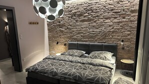 Ein Schlafzimmer mit Backsteinwand, einem Bett mit gemusterten Textilien, einer Hängelampe und einem kleinen runden Tisch.