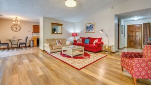 Sala de estar com sofá vermelho, mesa de centro branca e poltrona estampada. Há uma área de jantar com mesa e cadeiras, e uma cozinha com armários e espelho.