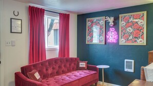 Un sofá rojo, una pequeña mesa redonda, un letrero de neón y cuadros con motivos florales sobre una pared oscura.