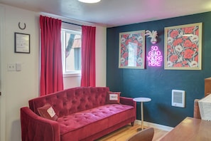 Ein rotes Sofa, ein kleiner runder Tisch, eine Neonreklame und gerahmte Blumenbilder an einer dunklen Wand.