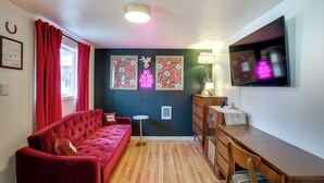 Una habitación con un sofá rojo, una mesa de comedor de madera, una mesita redonda pequeña, una lámpara y un televisor instalado en la pared.