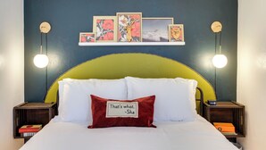 Una cama con ropa blanca, un almohadón rojo con texto y un cabecero verde.